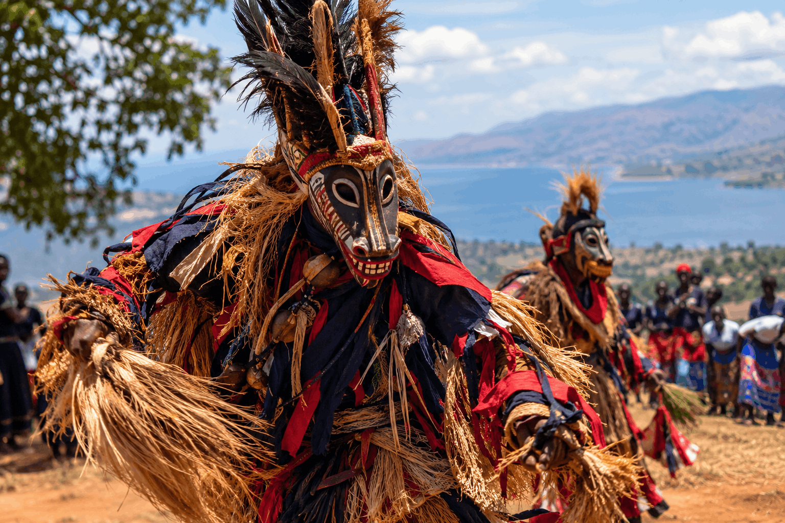 A masked Gule Wamkulu dancer in elaborate straw and feather costume performs in a Malawian cultural gathering, with other masked dancers, spectators, Lake Malawi and distant mountains in the background.