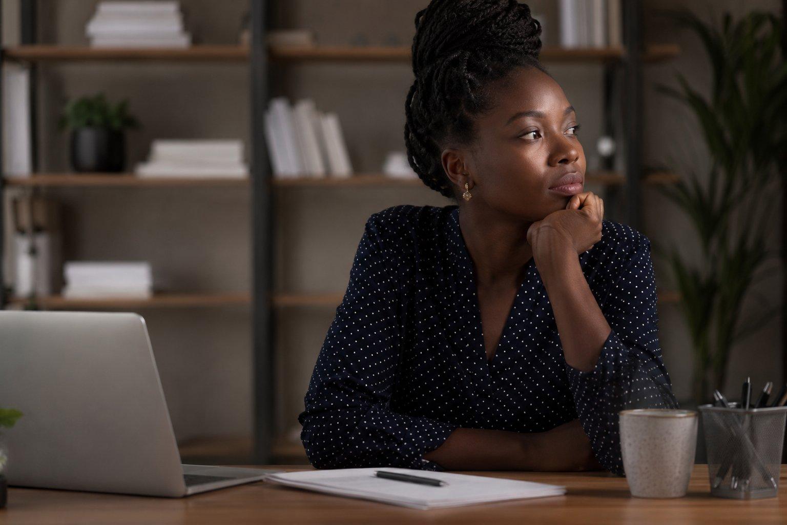 A thoughtful young African professional sits at her desk in a softly lit office, looking away from her laptop in quiet reflection. A notebook, pen, and coffee mug rest on the desk while blurred bookshelves and plants create a calm background, capturing a moment of self-reflection about confidence, achievement, and self-perception.