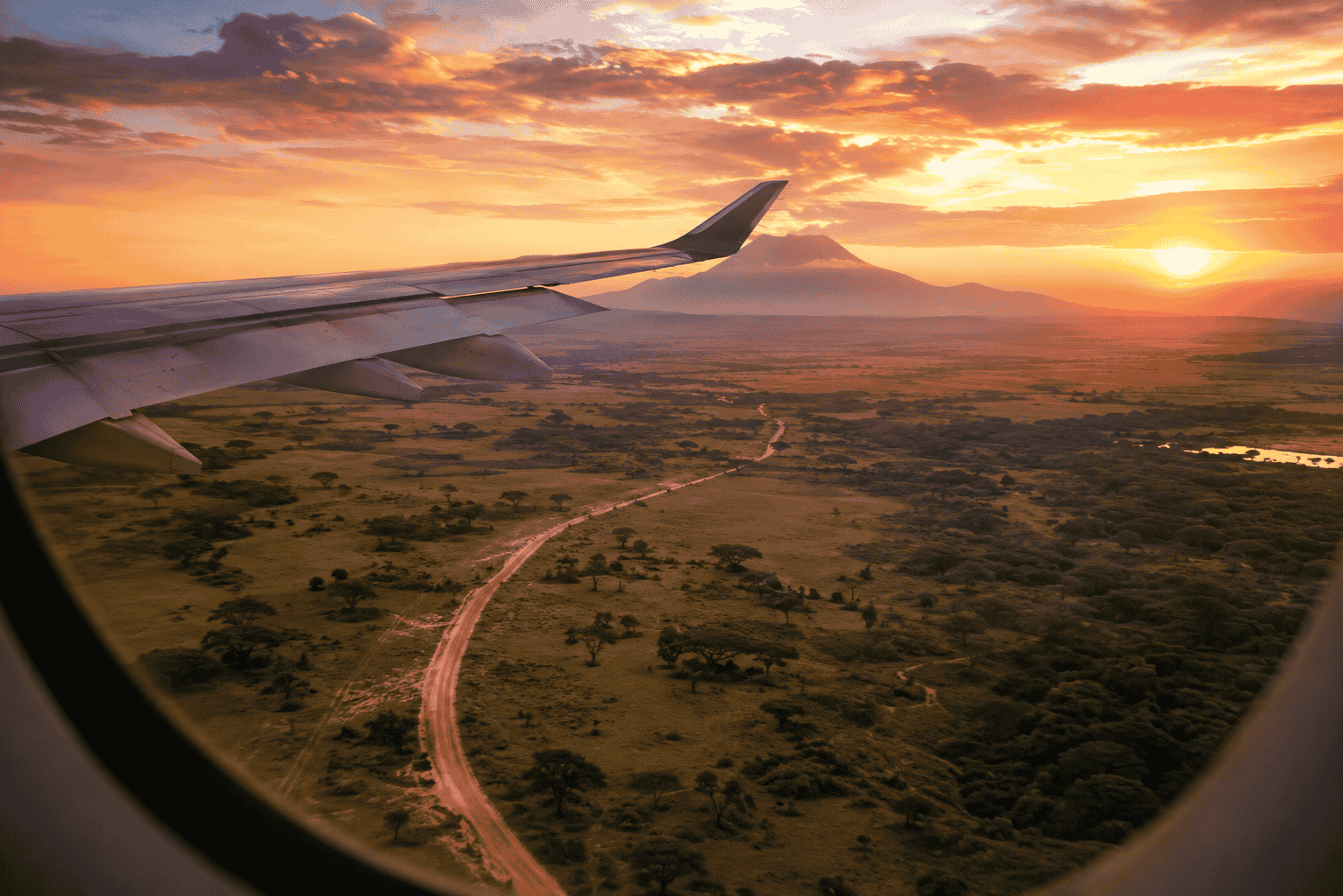 Aerial view from an airplane over Kenya’s savanna at sunset, with a winding road, distant mountain and warm golden landscape visible below.
