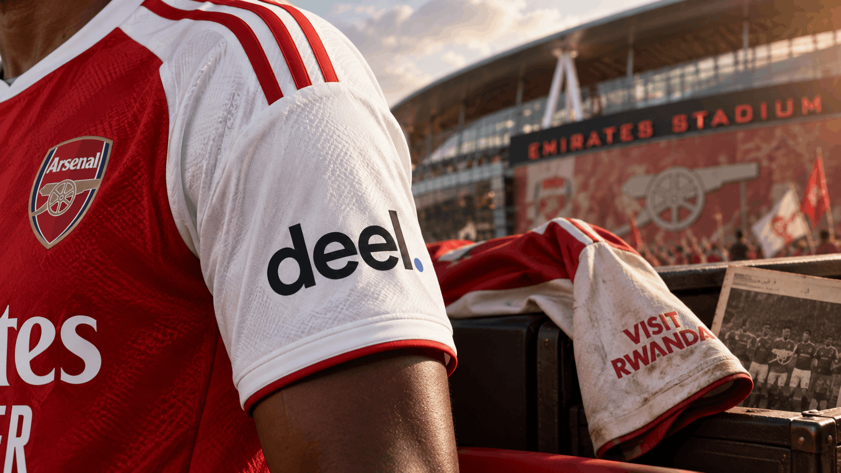 Close-up of an Arsenal shirt sleeve showing the Deel logo, with a faded Visit Rwanda sleeve from an old shirt draped nearby and Emirates Stadium in the background